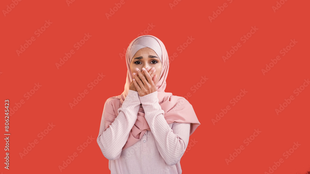 Fototapeta premium Shock expression. Fear emotion. Stressed scared sad overwhelmed woman in headscarf covering mouth with hand isolated on red empty space background.