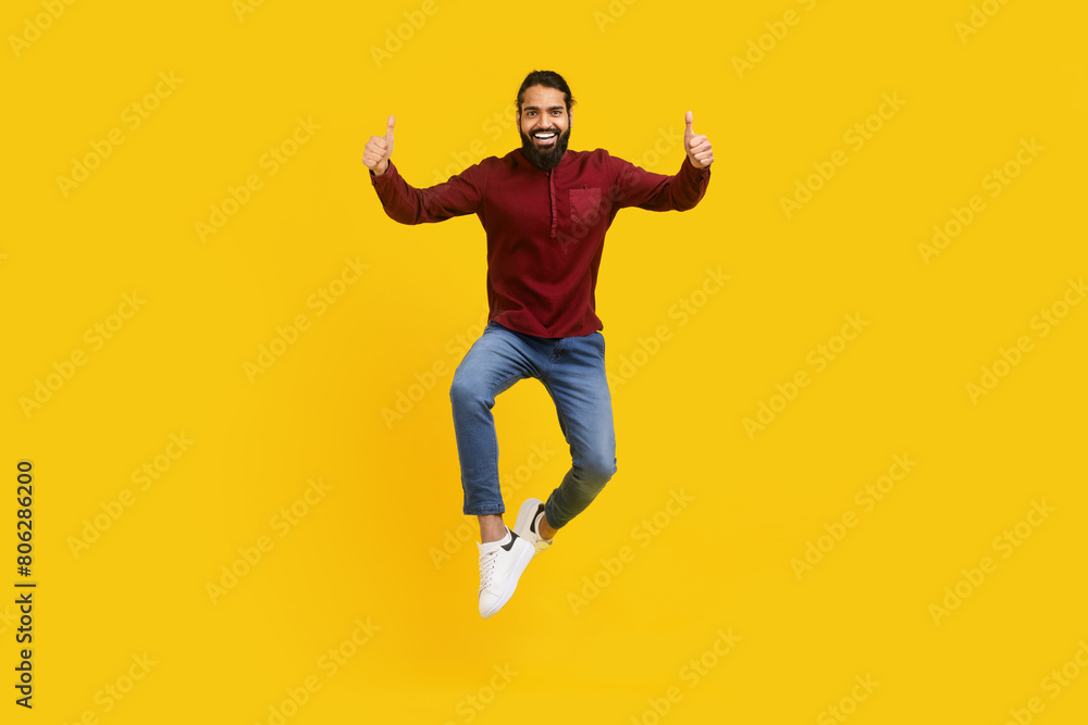 Obraz premium Indian man is captured mid-air, jumping energetically with his hands raised in thumb ups gesture, yellow studio background