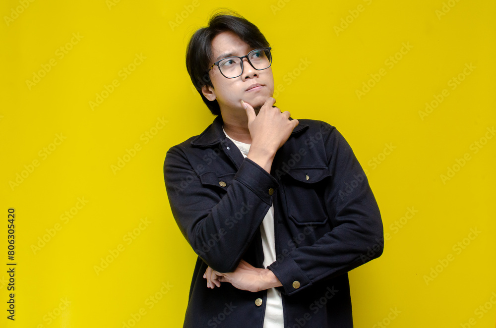Fototapeta premium man feeling uncertain and unsure. Thoughtful asian man thinking and wondering with hand touching skin as thinking gesture isolated over yellow background. 