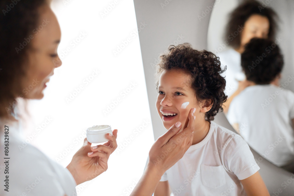 A young child, laughing with joy, receives gentle care from his mother as she applies moisturizer to his cheek. African American family standing close together in a well-lit bathroom A young child, laughing with joy, receives gentle care from his mother as she applies moisturizer to his cheek. African American family standing close together in a well-lit bathroom