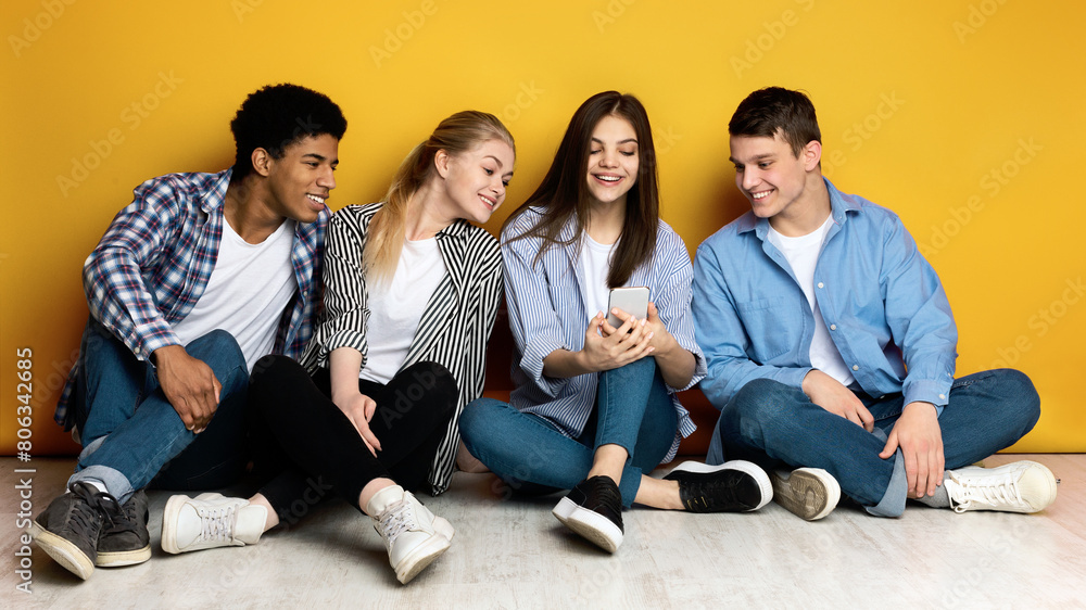 Four friends multiethnic teenagers are sitting closely together on the floor, leaning against a bright yellow wall. One of them is holding a smartphone, and they are all looking at the screen Four friends multiethnic teenagers are sitting closely together on the floor, leaning against a bright yellow wall. One of them is holding a smartphone, and they are all looking at the screen
