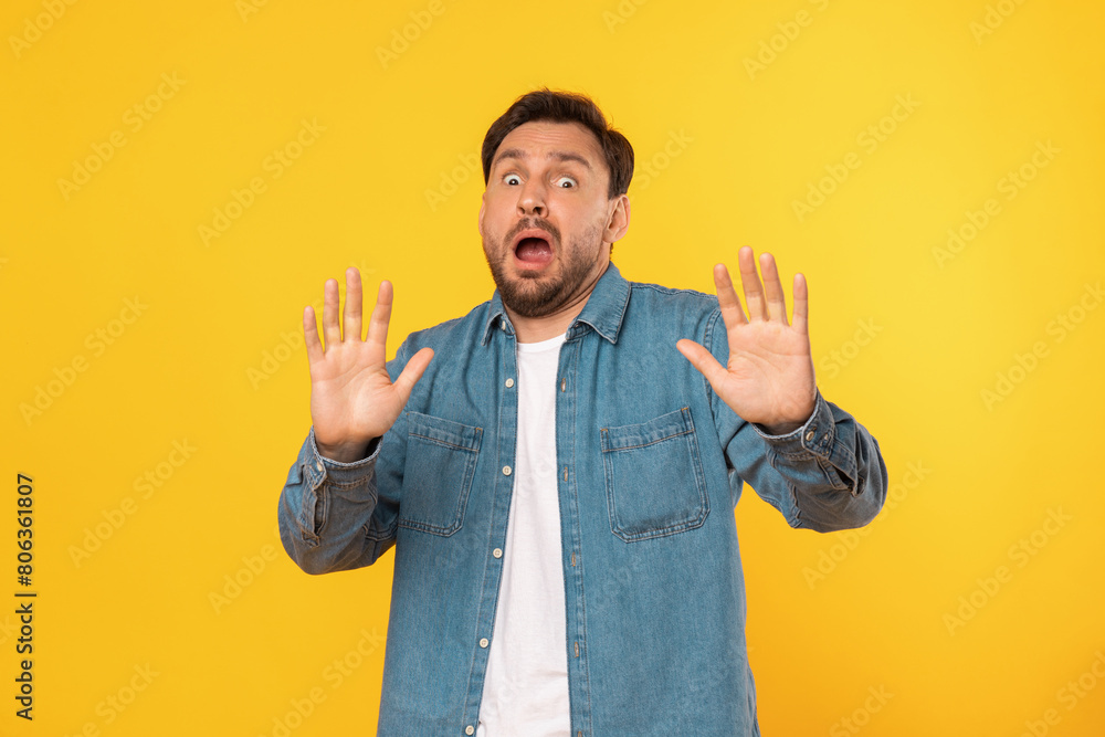 A young man with a beard stands against a bright yellow backdrop, showing a surprised expression with his mouth open and hands raised in a defensive or startled gesture. A young man with a beard stands against a bright yellow backdrop, showing a surprised expression with his mouth open and hands raised in a defensive or startled gesture.