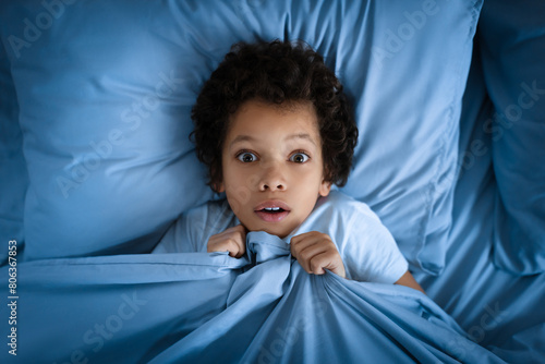 Принти на полотні African American young boy lies in bed with a surprised look on his face, clutching the blue bed covers up to his chin