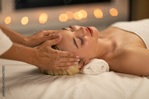 In a tranquil spa environment, a young woman with blonde hair experiences a soothing facial massage, radiating relaxation and peace. photo