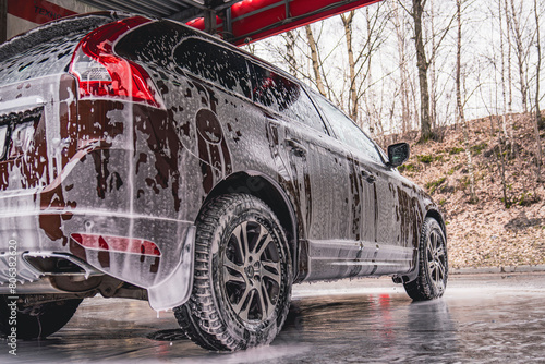 Vehicle covered in white soapy foam during regular carwash outdoors, auto getting professional wash with soap at washing center, cleaning car exterior surface at self-service station