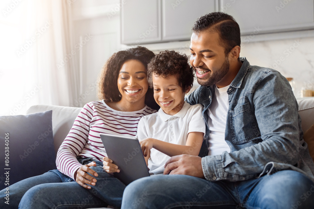 African American man, woman, and child are sitting together on a couch, engrossed in looking at a tablet. The man holds the device, while the woman and child lean in, focusing on the screen. African American man, woman, and child are sitting together on a couch, engrossed in looking at a tablet. The man holds the device, while the woman and child lean in, focusing on the screen.