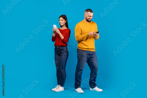 A man in a yellow sweater and jeans and a woman in a casual red top with dark denim jeans stand side by side, each absorbed in their own smartphone, blue background © Prostock-studio