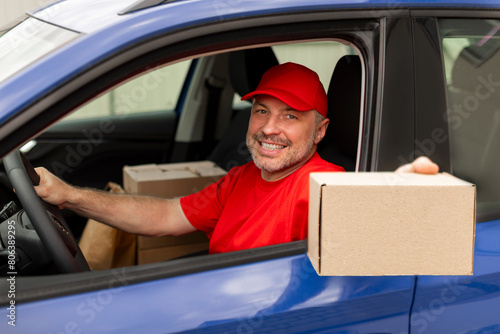 Happy middle aged courier man sitting in car, delivering package and giving it to client, stretching hand to camera