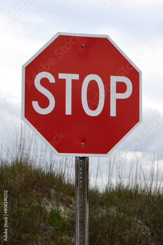 A bright red stop sign with the dunes of the beach and grey cloudy sky in the background.