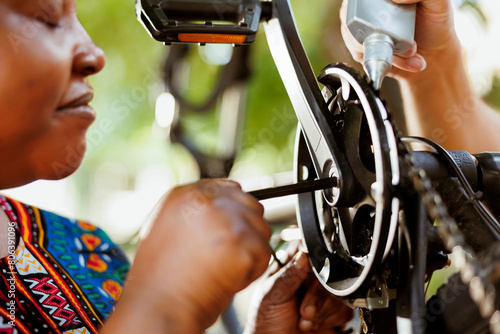 Close up of female african american using allen key to repair damaged bicycle crank-arm. Outdoor detailed shot of two people repairing and greasing bike chain and pedals for secure outdoor cycling. photo