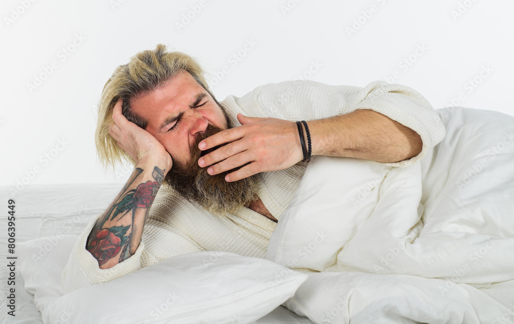 Bearded man yawning lying on soft white pillow. Handsome man in bathrobe yawning waking up in cozy bed at home in morning. Sleepy man in bed at bedroom yawning covering mouth with palm. Yawn and nap.