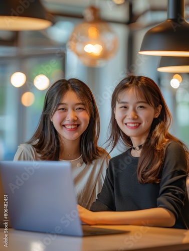 Two smiling Asian women sit in front of their laptops, exuding happiness and focus in a modern, brightly lit corporate office setting Cinematic Mood and tone.