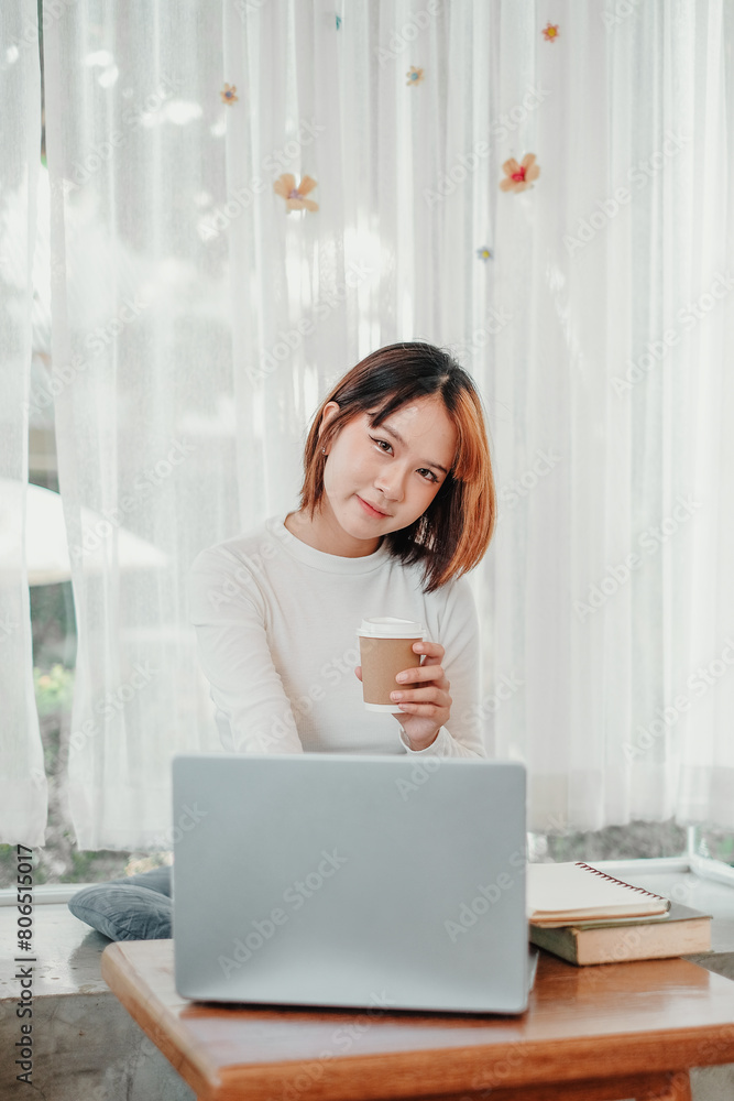 Young woman casually holding a coffee cup, taking a moment break from work, seated in front of her laptop in a bright, airy room. Young woman casually holding a coffee cup, taking a moment break from work, seated in front of her laptop in a bright, airy room.