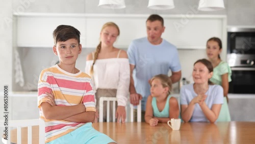 Offended pre-teen boy standing with arms crossed over chest at kitchen on background of his big family. High quality 4k footage photo