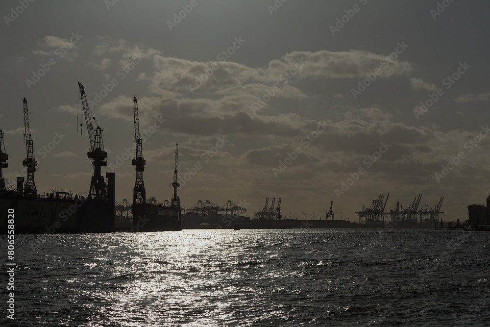 Port of Hamburg at sunset with dark backdrop of container cranes and sunlight at Elbe river.