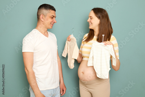 Young married pregnant family preparing for childbirth future mother showing to dad new tiny bodysuits buying for expecting kid standing isolated over light green background