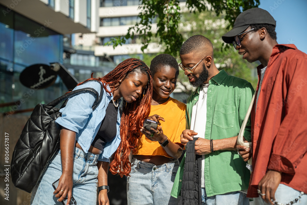 Interested stylish group of african american young people students looking at smartphone while walking in street. Happy black woman showing something to joyful friends in cellphone have fun outdoors. Interested stylish group of african american young people students looking at smartphone while walking in street. Happy black woman showing something to joyful friends in cellphone have fun outdoors.