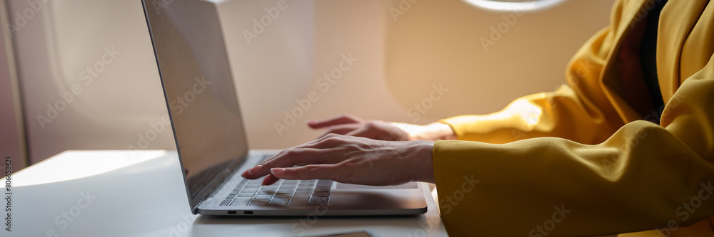 Fototapeta premium Young Asian business woman in yellow suit uses laptop sitting near window on airplane to do online financial business work Contact during flight, travel and business tourism concept.