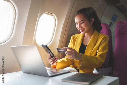 Young Asian businesswoman holding credit card and smartphone in online shopping using website on laptop sitting near window on airplane during flight, travel and business tourism concept.