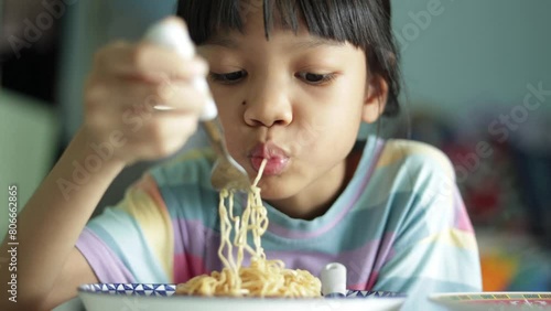 Cute Asian kid girl Standing and eating deliciously fried yellow noodles on a plate with a fork. She used a fork to put it in her mouth and chewed quickly because of hunger.