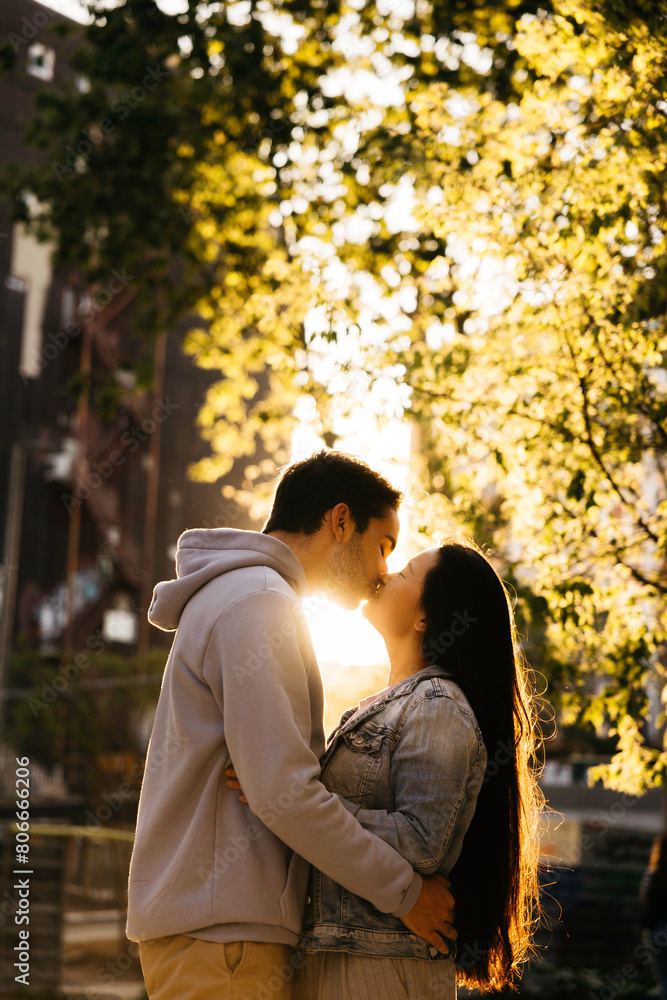 Young interracial couple kissing outdoors sorrounded by trees on the street of a city.