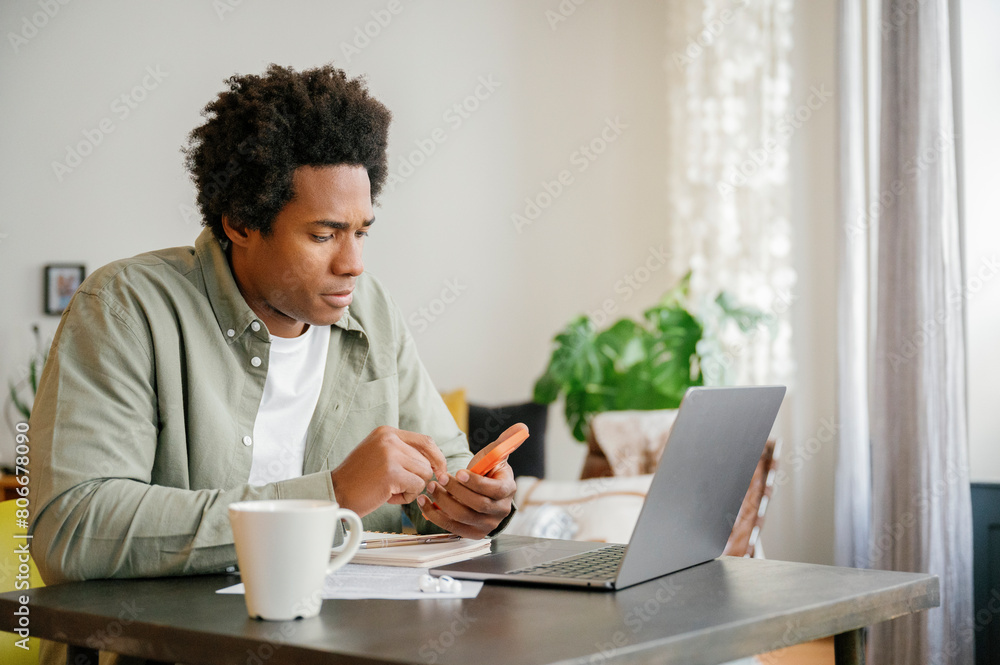 Businessman with Afro hairstyle using smart phone sitting with laptop working at home Businessman with Afro hairstyle using smart phone sitting with laptop working at home