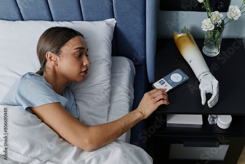 Young woman turning off alarm on smart phone by bionic arm in bedroom photo