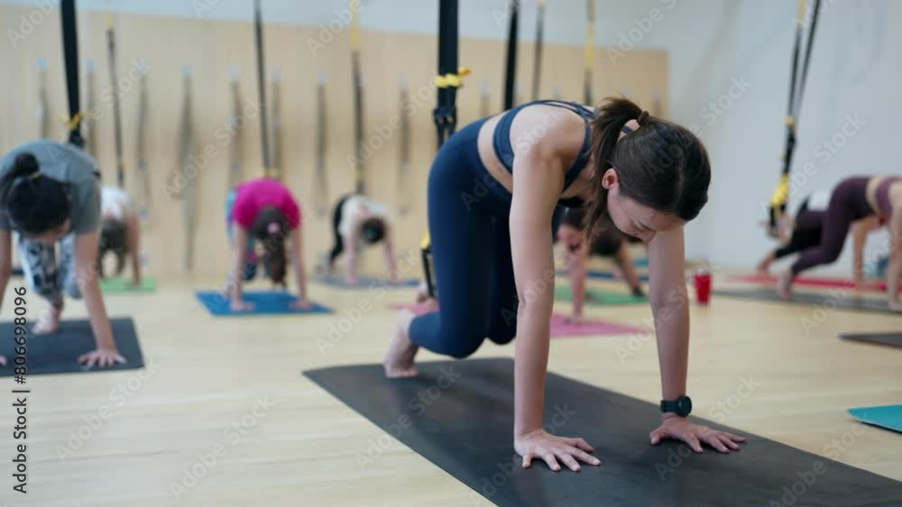A woman is doing yoga on a mat in front of a wall. There are several other people in the background doing yoga as well A woman is doing yoga on a mat in front of a wall. There are several other people in the background doing yoga as well