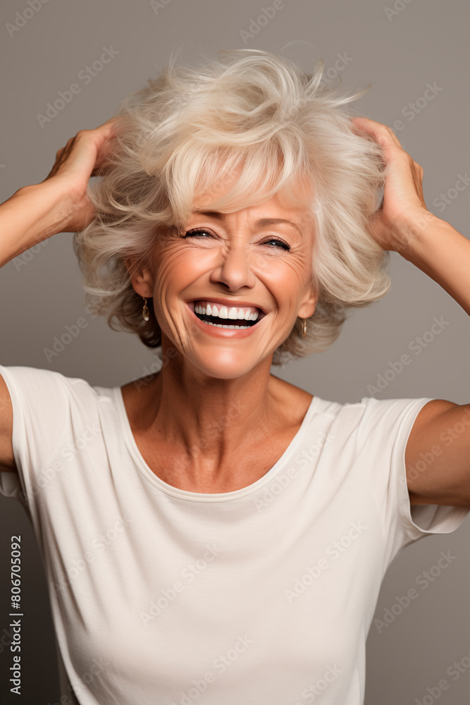 An older woman with blonde hair joyfully stretches her hands upwards, her face lit by a radiant smile, expressing happiness and vitality in a casual yet stylish portrait. An older woman with blonde hair joyfully stretches her hands upwards, her face lit by a radiant smile, expressing happiness and vitality in a casual yet stylish portrait.