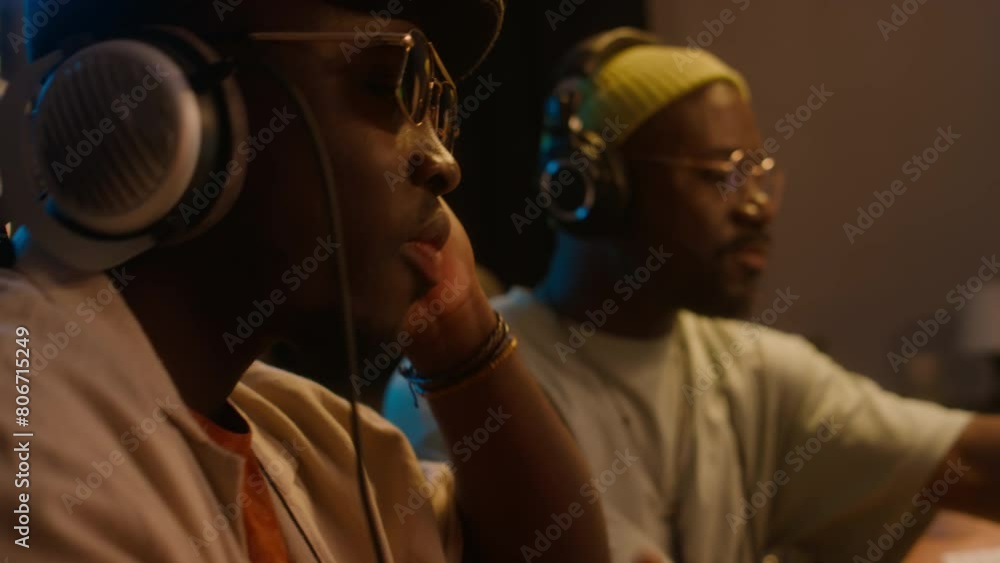 Medium side close-up of two African American electronic musicians sitting in recording studio, listening to new hip-hop music track in headphones, enjoying it and rocking to rhythmMedium side close-up