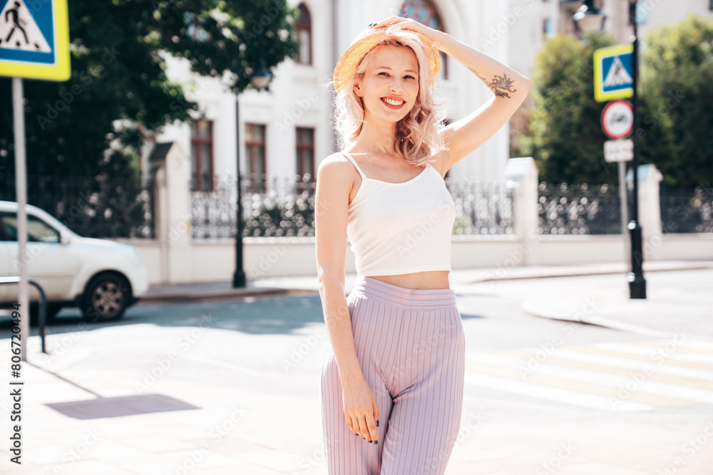 Young beautiful smiling blond hipster woman in trendy summer clothes. Carefree female posing in the street at sunny day. Positive model outdoors at sunset. Cheerful and happy in hat