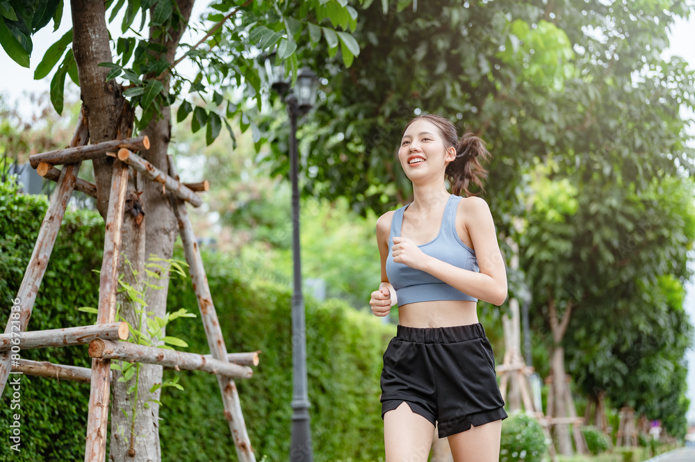 weight, jogger, fit, fitness, exercising, marathon, run, runner, jogging, vitality. A woman is running in a park with trees in the background. She is smiling and she is enjoying her run. weight, jogger, fit, fitness, exercising, marathon, run, runner, jogging, vitality. A woman is running in a park with trees in the background. She is smiling and she is enjoying her run.