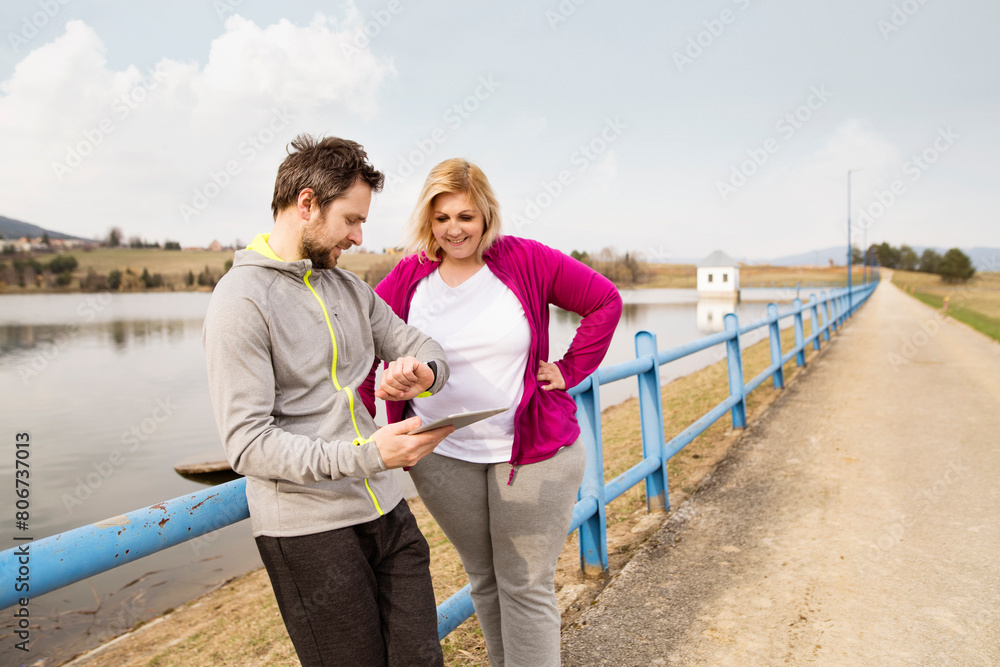 Overweight woman resting after run, personal trainer checking her profile, performance on tablet. Exercising outdoors for people with obesity, support from fitness coach. Overweight woman resting after run, personal trainer checking her profile, performance on tablet. Exercising outdoors for people with obesity, support from fitness coach.
