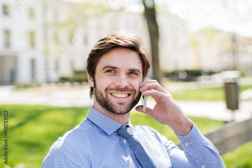 Handsome businessman phone calling on smartphone, sitting on bench in city. Working remotely, waiting for business meeting. Manager smiling, outdoor in urban setting