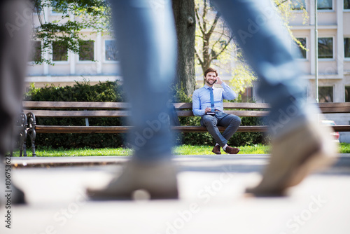 Handsome businessman phone calling on smartphone, sitting on city bench, writing notes. Working remotely, waiting for business meeting. Manager smiling, outdoor in urban setting