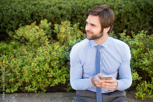 Handsome businessman holding smartphone sitting on bench in city park. Working remotely, waiting for business meeting. Manager smiling, outdoor in urban setting