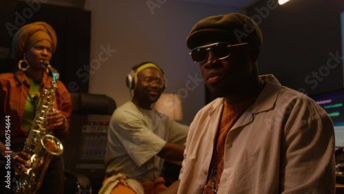 Medium of African American male musician in cap and sunglasses playing piano keyboard, woman performing on sax, audio engineer in headphones manning mixing board, while recording music in studio