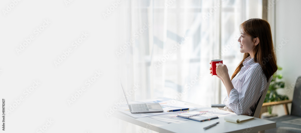 A woman is sitting at a desk with a red mug in front of her. She is smiling and she is enjoying her coffee. The scene suggests a relaxed and comfortable atmosphere