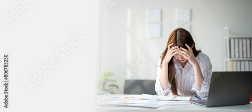 A woman is sitting at a desk with a laptop and a stack of papers. She is looking at the laptop and she is in a state of distress photo