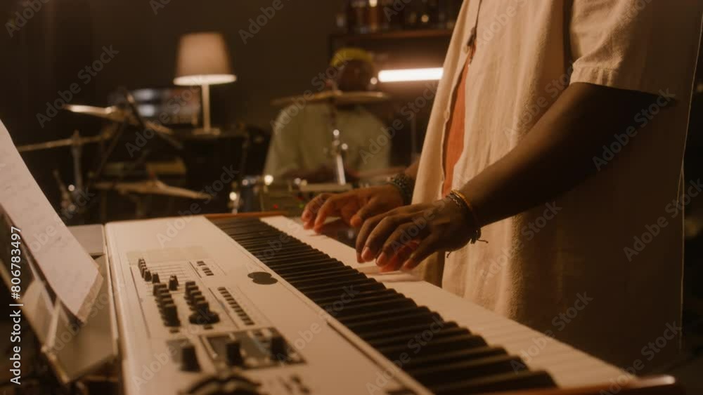 Medium tilting of young male African American musician in cap and sunglasses playing keyboards, male bandmate sitting on drums, while recording new music or rehearsing together in studio