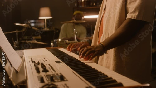 Medium tilting of young male African American musician in cap and sunglasses playing keyboards, male bandmate sitting on drums, while recording new music or rehearsing together in studio photo