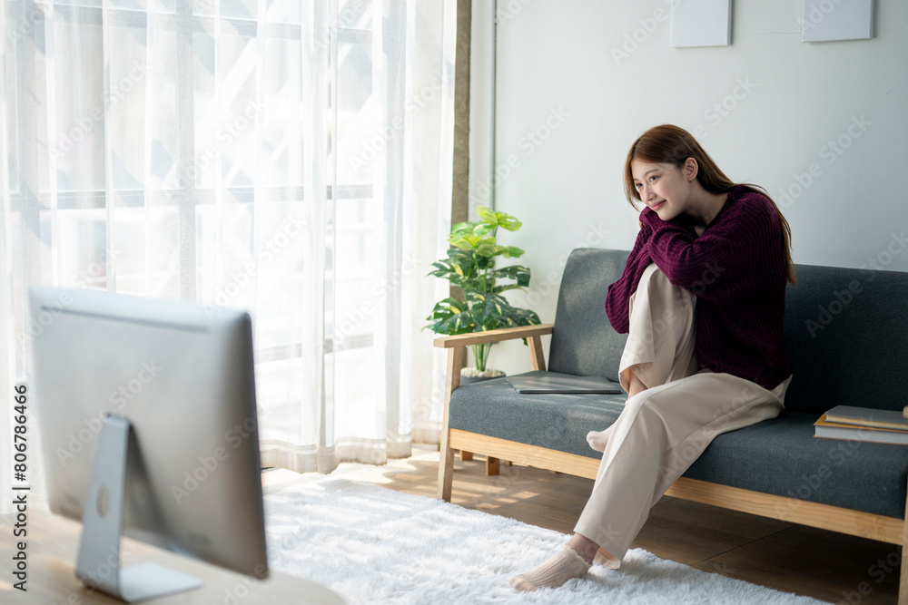A woman is sitting on a couch in a living room, watching television. She is wearing a purple sweater and has her legs crossed. The room has a TV on the left side and a potted plant on the right side