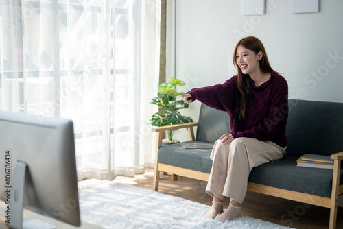 A woman is sitting on a couch in a living room, pointing at something on the television. She is smiling and she is enjoying herself. The room is decorated with a potted plant and a laptop