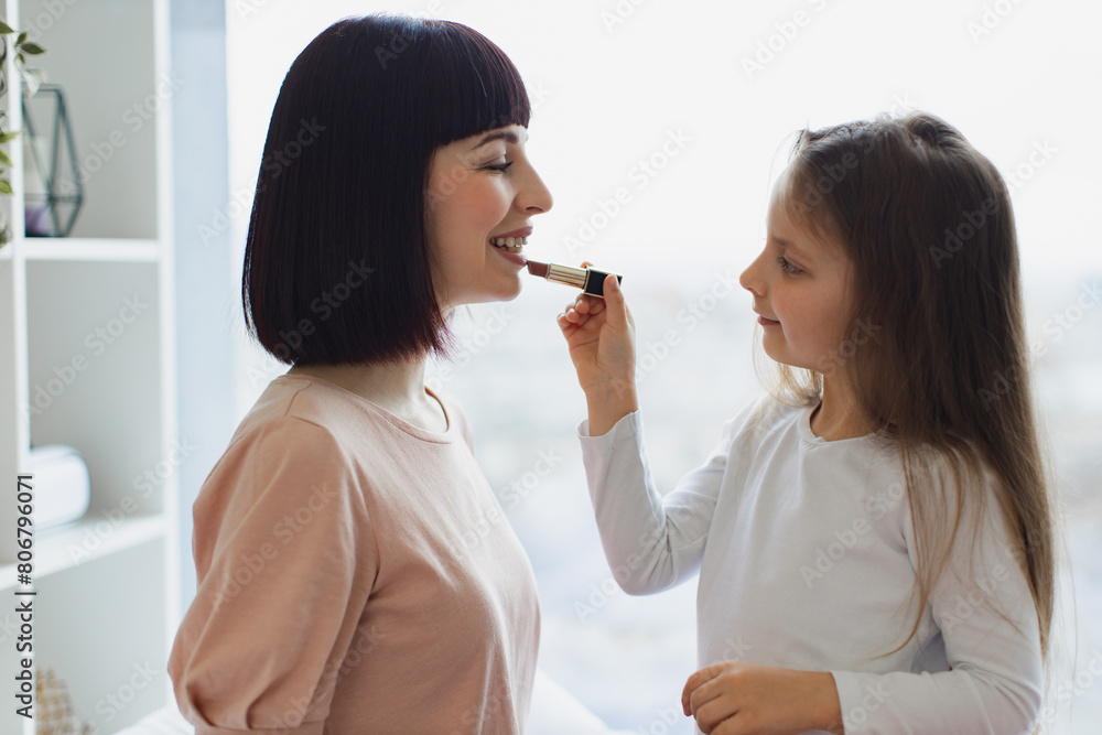 The daughter paints lips to her beautiful mommy. Little girl doing makeup to mom sitting at bedroom. Happy Mother and daughter having fun together at weekend. Happy loving family. The daughter paints lips to her beautiful mommy. Little girl doing makeup to mom sitting at bedroom. Happy Mother and daughter having fun together at weekend. Happy loving family.
