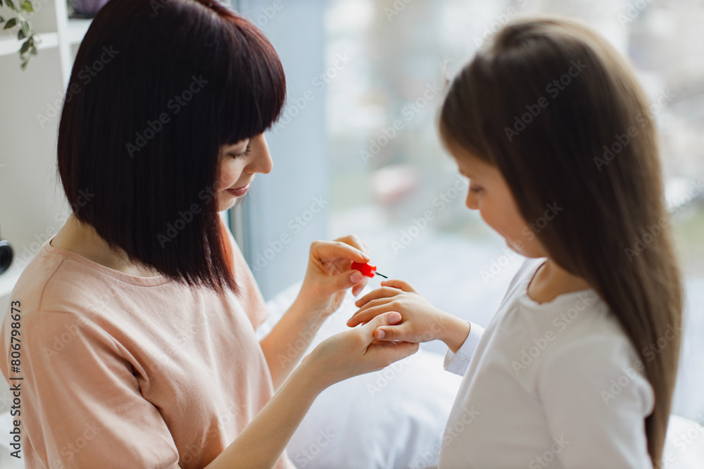 Young happy woman paints her little girl nails while sitting on the bed near panoramic window. Caucasian mother and daughter doing manicure on weekend at home. Young happy woman paints her little girl nails while sitting on the bed near panoramic window. Caucasian mother and daughter doing manicure on weekend at home.