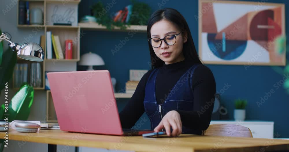 Cute Asian woman sitting in front of laptop. Focused on working remotely while hearing phone ringing. Picking up smartphone. Beginning conversation with co-worker or relative. Distracted girl. Cute Asian woman sitting in front of laptop. Focused on working remotely while hearing phone ringing. Picking up smartphone. Beginning conversation with co-worker or relative. Distracted girl.