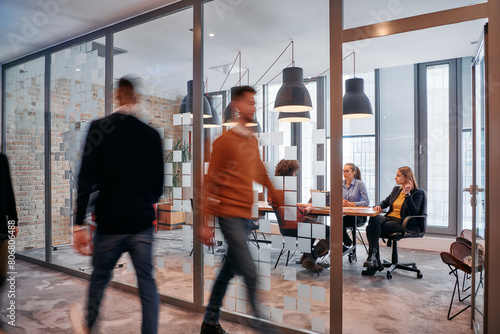 In the dynamic hustle and bustle of a business environment, a group of young business professionals walk down the corridor next to their office, while their colleagues collaborate inside, symbolizing