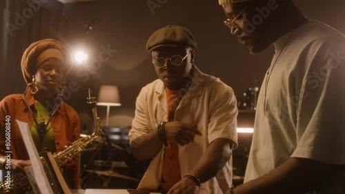 Medium of two African American male musicians standing at electric piano in studio, playing various chords on keyboards, discussing new song, smiling woman in turban, with saxophone listeningMedium of