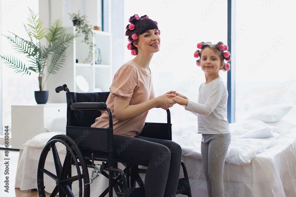 Obraz premium Happy Caucasian woman and daughter doing hair curling. Young mother in wheelchair holds hands of her little daughter on weekend morning in bedroom.