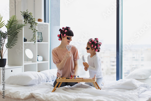 Cheerful mother and daughter singing using combs as microphones during beauty treatments at home on bed. Caucasian young woman and little girl curling hair and applying eye patches.